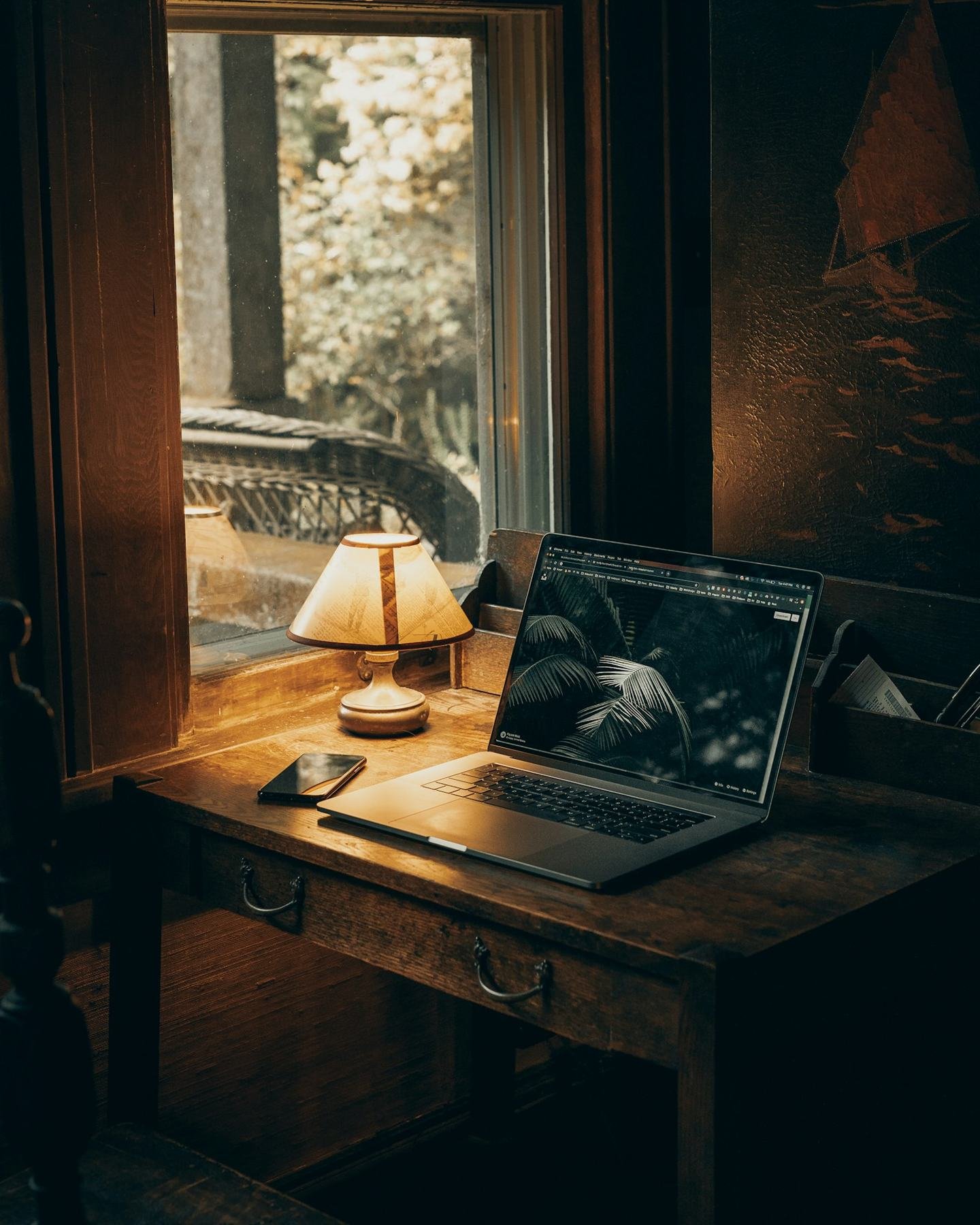A laptop on a wooden desk by a window in warm evening light